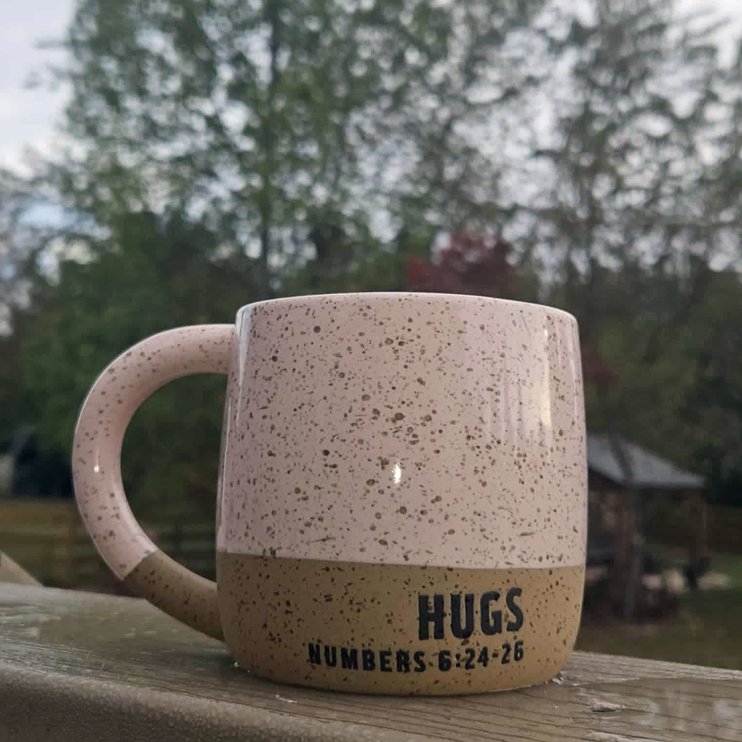 Speckled ceramic mug with 'HUGS NUMBERS 6:24-26' text on a wooden surface outdoors.