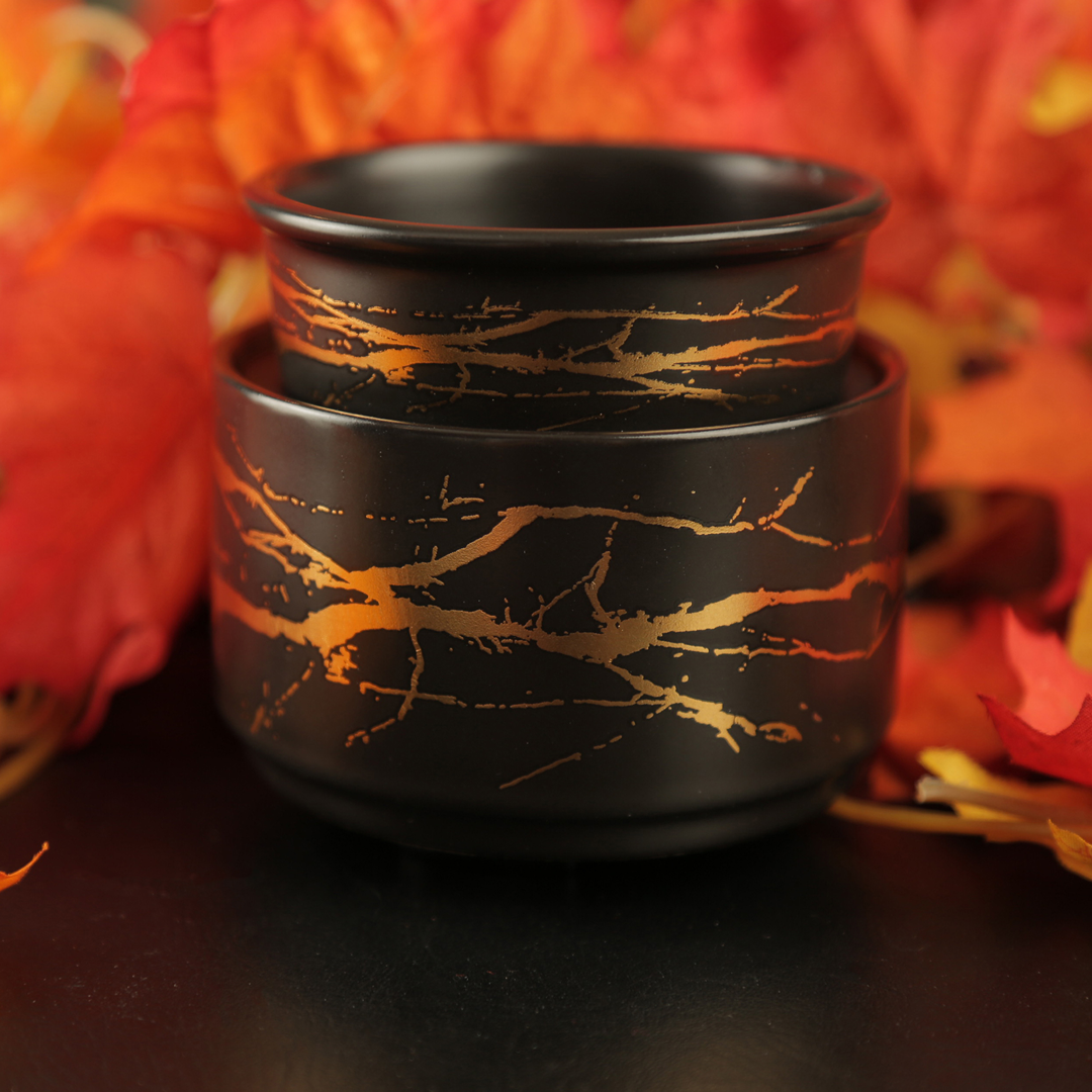 Black ceramic wax warmer with gold branch-like patterns on a dark background with red leaves.