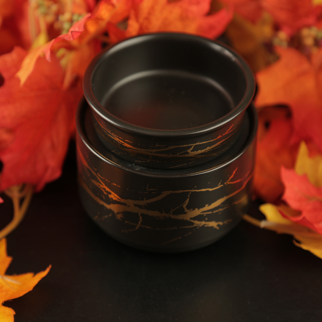 Black ceramic wax warmer with gold leaf design on a black background with autumn leaves.
