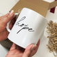 A white ceramic mug with the word 'hope' written in black cursive script, held by two hands in front of a brown cardboard box with shredded paper filler.
