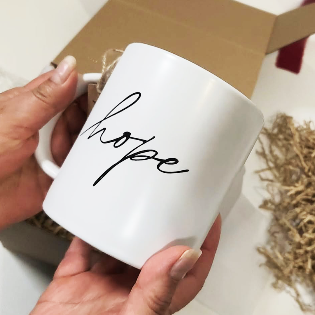 A white ceramic mug with the word 'hope' written in black cursive script, held by two hands in front of a brown cardboard box with shredded paper filler.