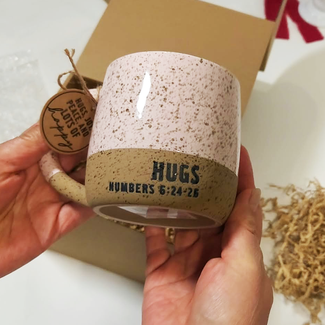 A person holds a speckled ceramic mug with a brown band printed with the words 'HUGS NUMBERS 5:24-26'. The mug has a light pink upper half and a beige lower half with brown speckles. The Cozy Sips Gift Box.