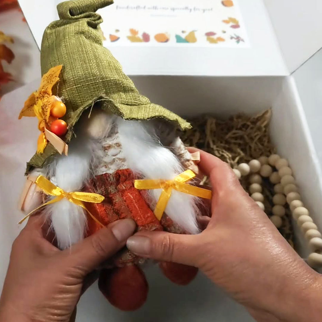 A small autumn-themed girl gnome figurine with a green burlap hat, white fluffy pony tails, and red patterned clothing, inside Gathered in Gratitude Thanksgiving gift box