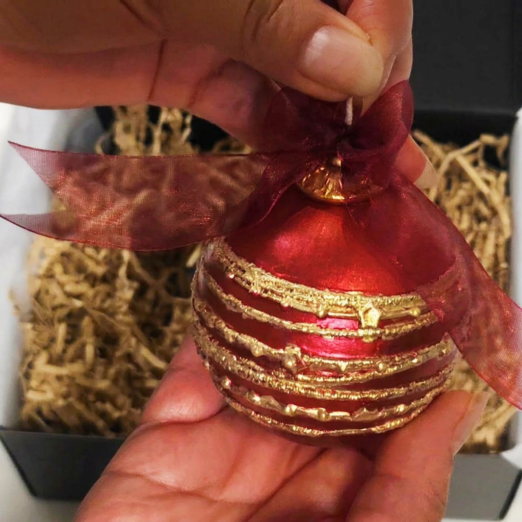 A red and gold striped Christmas ornament held in a person's hands, with a sheer red ribbon bow at the top, shown in front of a gift box filled with shredded paper.