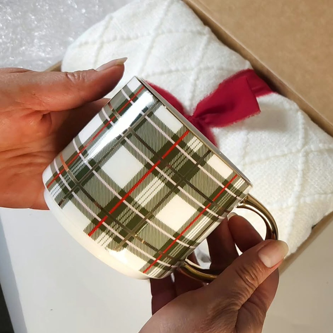 One green plaid ceramic coffee mug with a green, white, and red plaid pattern and a gold-toned handle, held by two hands next to a white quilted blanket with a red ribbon inside a gift box.