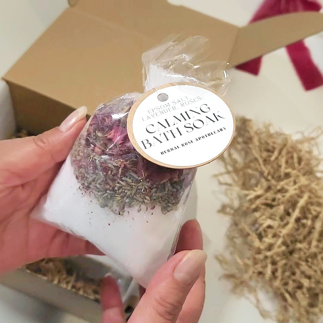 A hand holds a small clear bag of calming bath soak with Epsom Salt, dried flowers, and herbs, attached to a round label. The Cozy Moments Gift Box.
