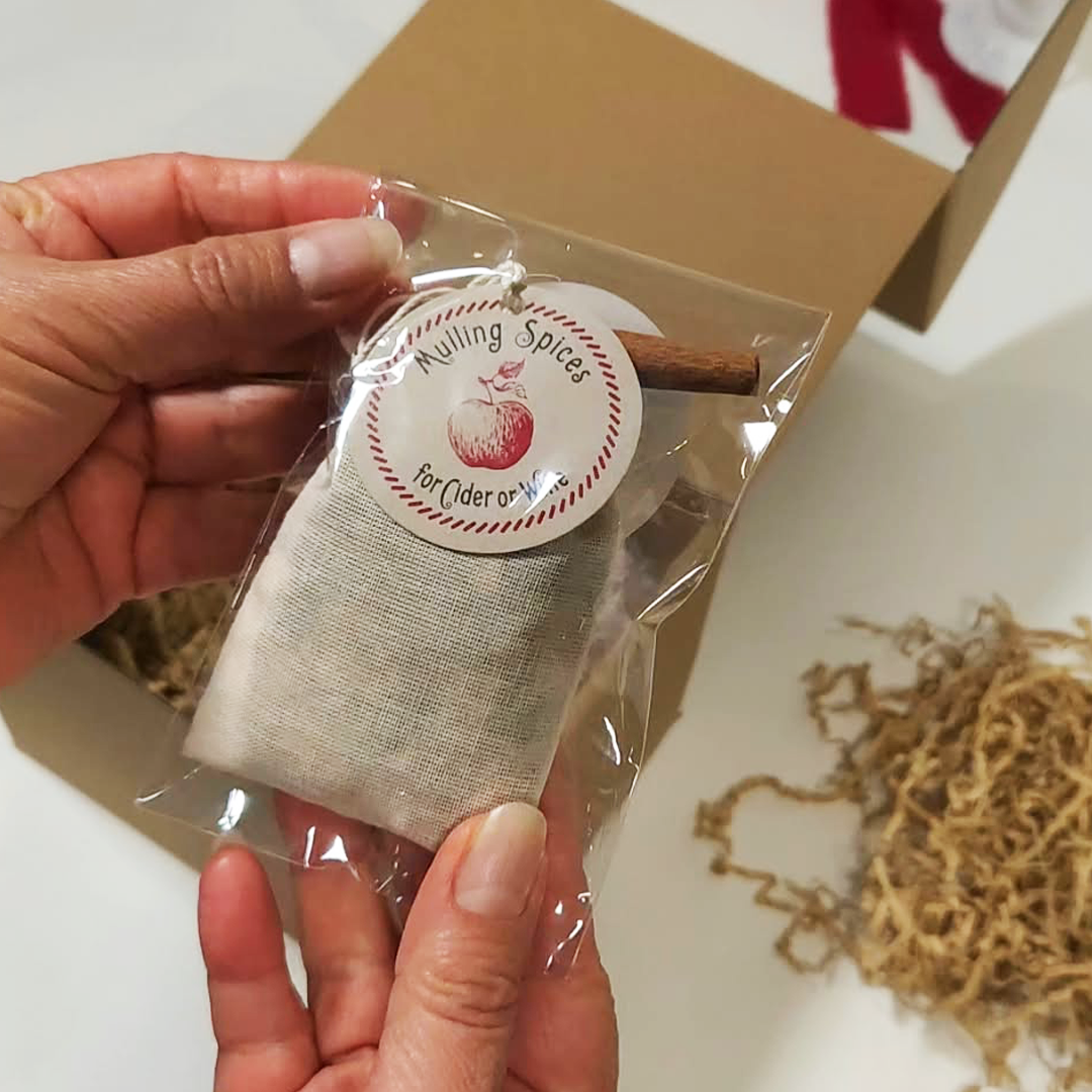 A hand holding a packet of mulling spices for cider or wine, with a round white label that reads Mulling Spices and a cinnamon stick visible. The Cozy Sips Gift Box.