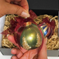 A hand holds a Green Scented Wax Baubles Ornament with a burgundy ribbon bow, with textured gold tree designs on the lower half, with other ornaments in a gift box behind it.