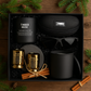 Gift set with black mug, candles, and sunglasses on a wooden surface with pine branches.