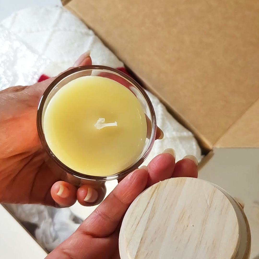 A hand holds a small clear 4oz glass candle jar filled with pale yellow wax, with a white wick visible. A light wood lid is placed beside a Christmas gift box.