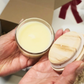 A hand holds a small hand-poured 4oz candle with a wooden lid. The Cozy Moments Gift Box.