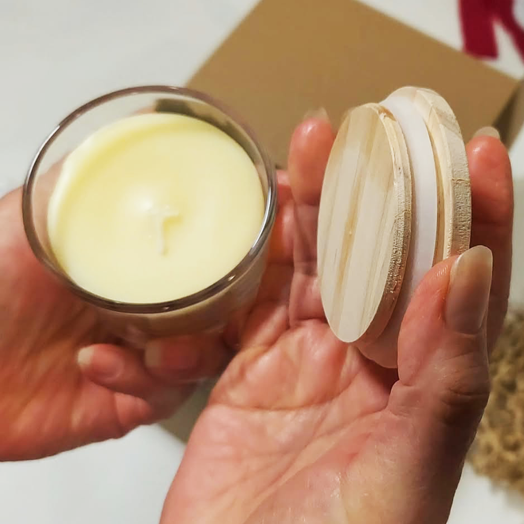 A hand holds a small hand-poured candle next to a round wooden lid. The Cozy Sips Gift Box.
