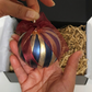 A hand holds a striped Blue Scented Wax Baubles Ornament with metallic blue, gold, and bronze bands tied with a burgundy ribbon, with a black gift box and shredded paper visible in the background.