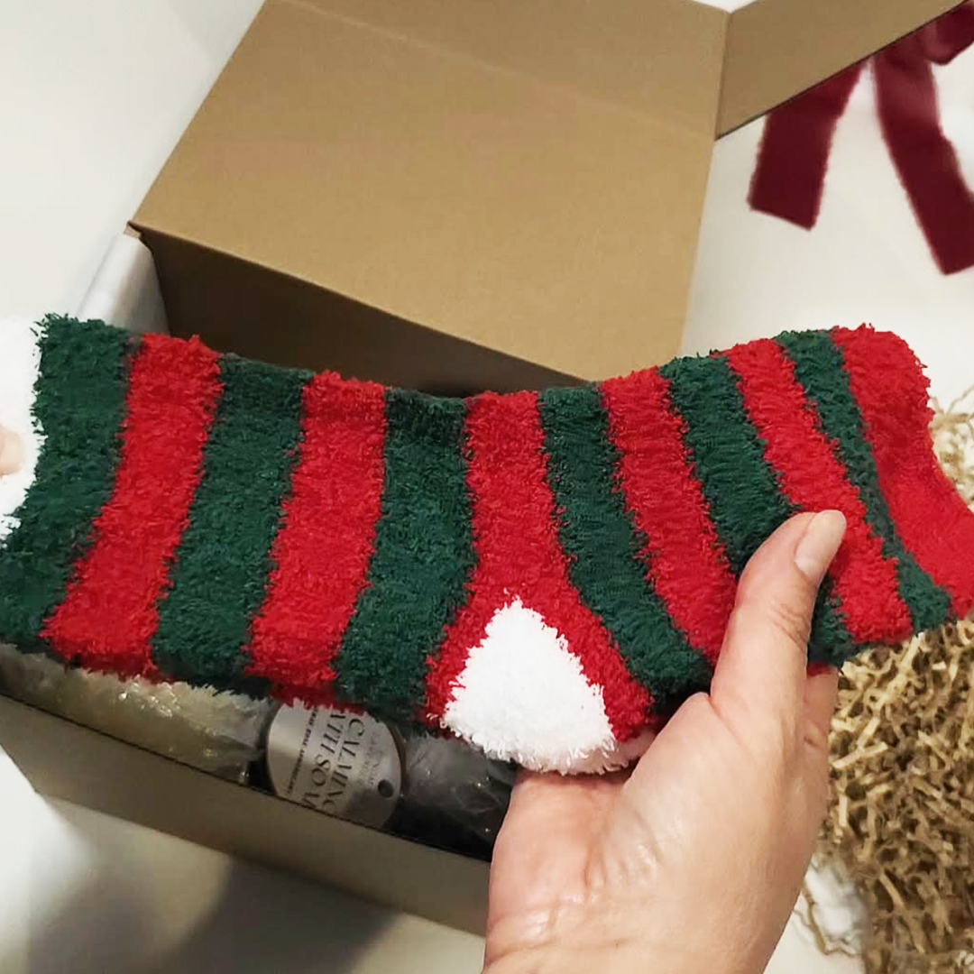 A hand holding a Holiday Fuzzy Fluffy Socks with red and green stripes and a white cuff. The Cozy Moments Gift Box