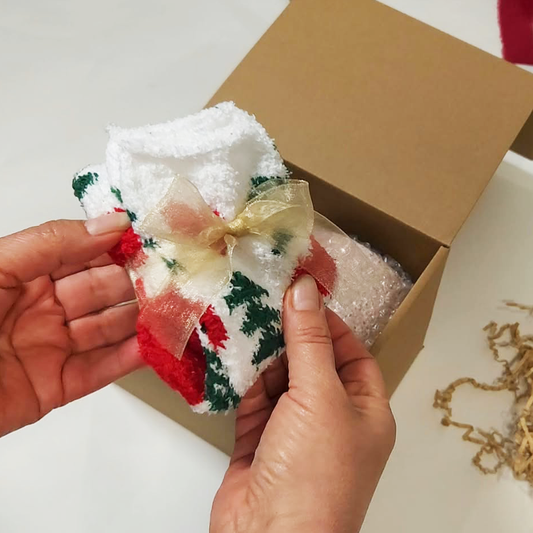 A person's hands holding a pair of holiday fuzzy fluffy socks with red and green holiday patterns and a sheer gold ribbon bow. The Cozy Sips Gift Box.