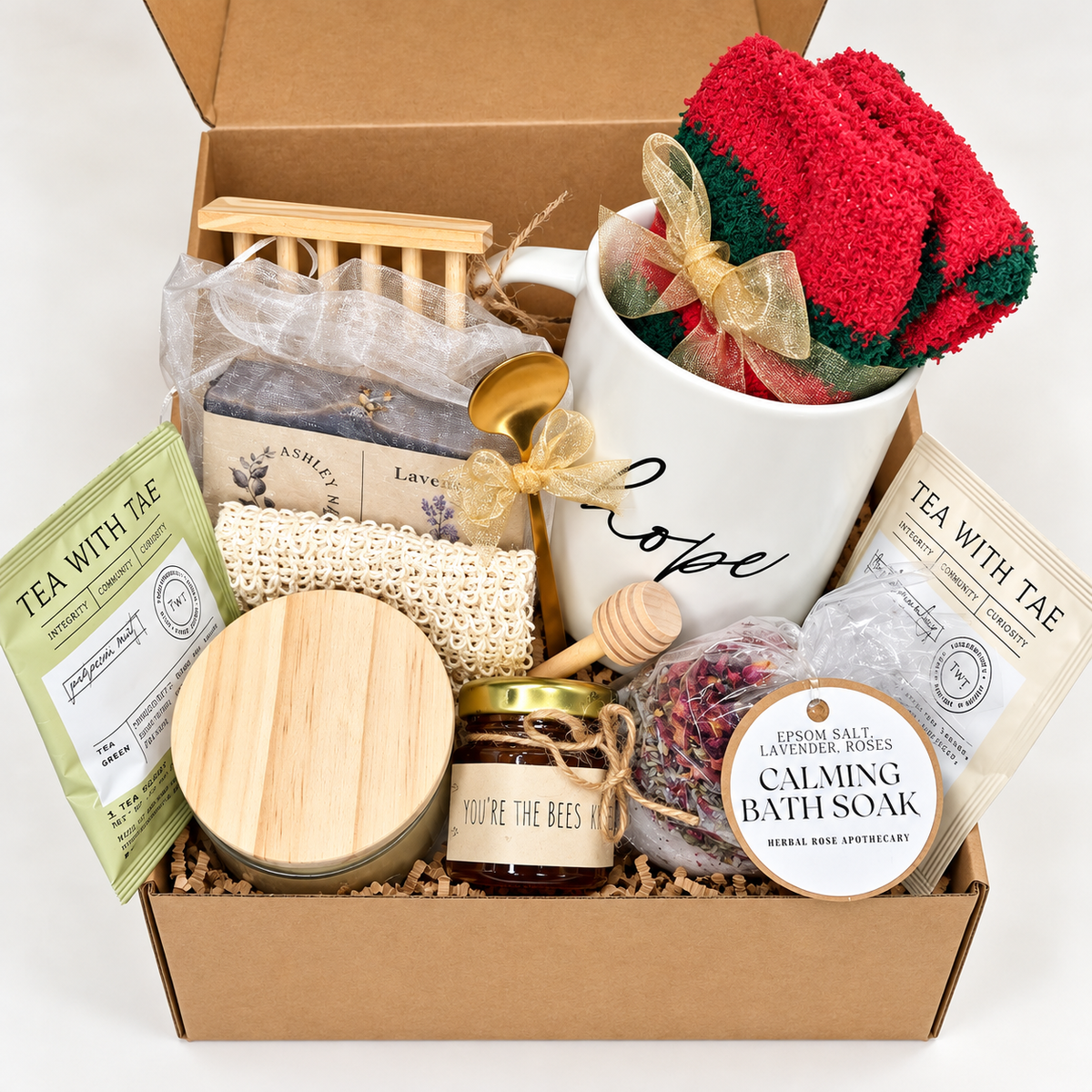 The Cozy Moments Gift Box combines bath and body treats with cozy comforts and tea-time favorites. It includes a herbal bath soak featuring Epsom salt, lavender, and roses, lavender soap, fluffy holiday socks, mini honey with a dipper, a gold coffee teaspoon, a thick glass 4 oz candle, a Hope mug, an exfoliating soap pouch, a bamboo wood soap holder, and two flavorful teas.