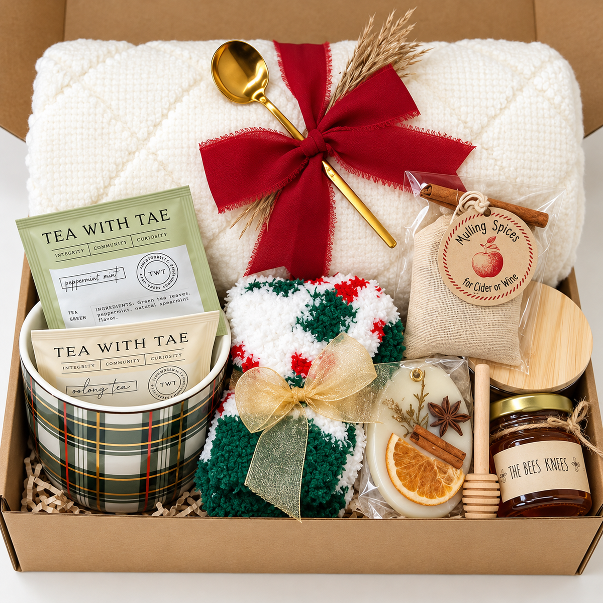 The Cozy Christmas Gift Box_ includes a Throw Blanket, a Mini Honey w/Deeper, a Holiday Fuzzy Fluffy Socks, a 4oz candle, a Green Plaid Coffee Mug, a Gold Coffee Teaspoon and packaged tea bags and accessories. Arranged on a rustic wooden table with festive decorations.