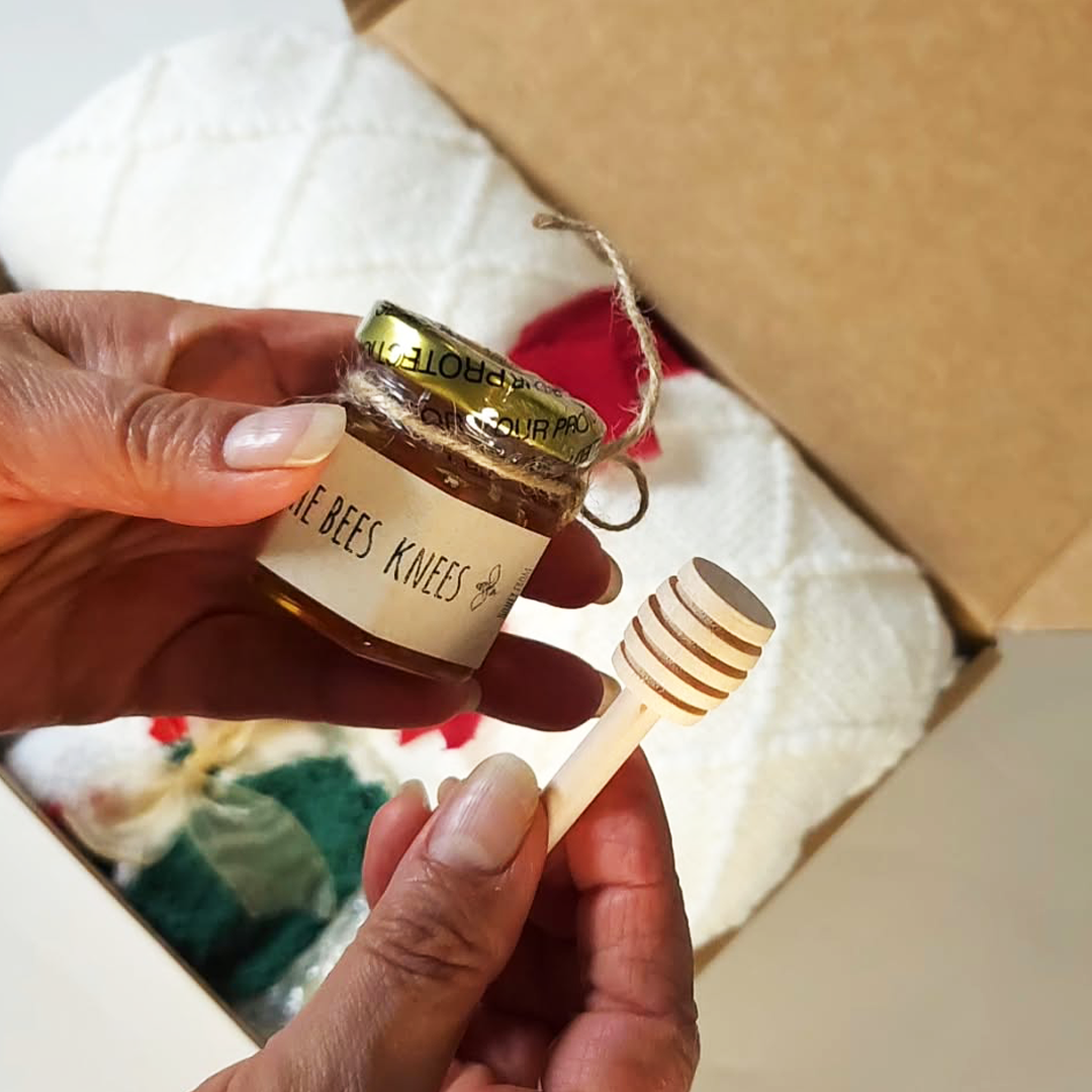 A person holds a small glass jar of honey with a gold foil lid and a wooden honey dipper in front of an open Christmas gift box.