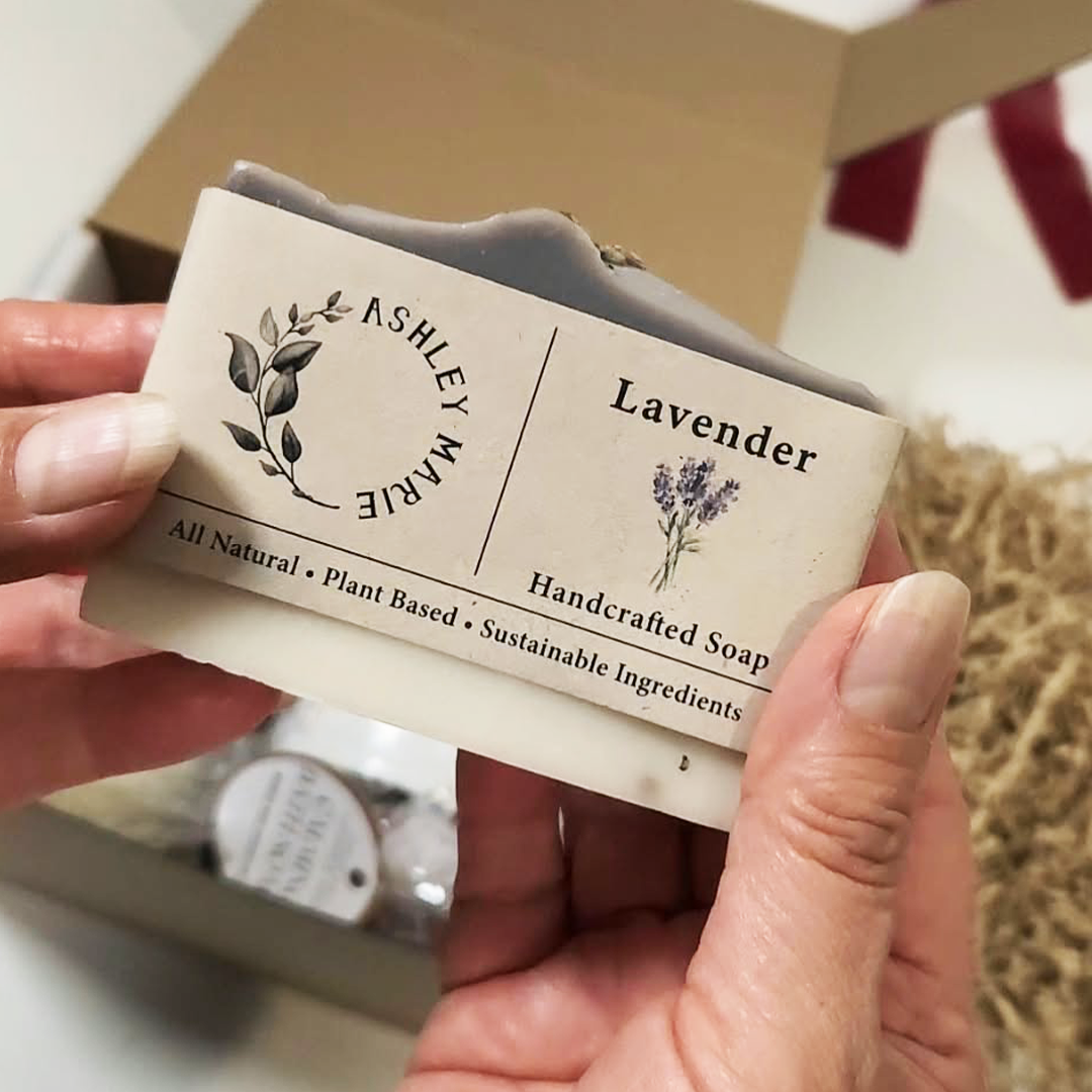 A hand holding a soap bar of Ashley Marie Lavender Handcrafted Soap, showing the cream-colored packaging and lavender illustration, with The Cozy Moments Gift Box.