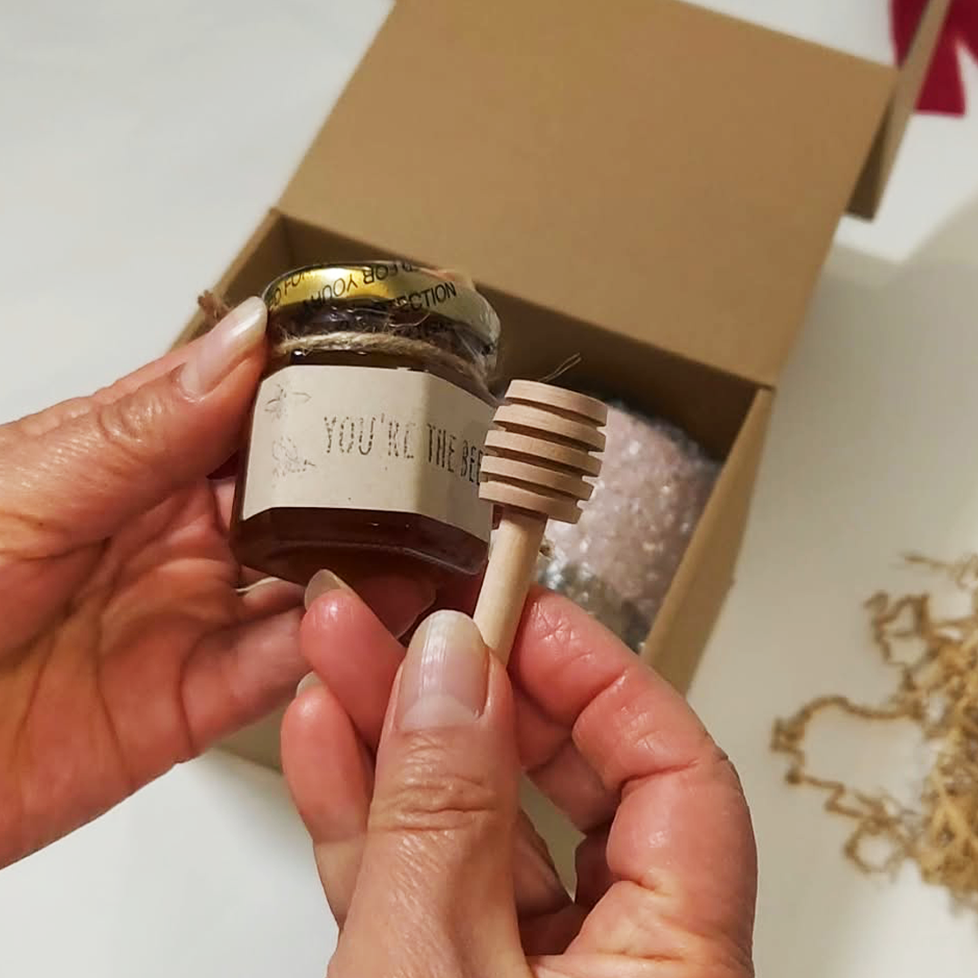 A person's hands hold a small glass jar of honey with a wooden honey dipper. The Cozy Sips Gift Box.