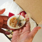 A hand holds a small oval white ornament decorated with dried orange slices, cinnamon sticks, star anise, and green sprigs, with a sheer beige ribbon loop. An open Christmas gift box with a cozy blanket inside the box.