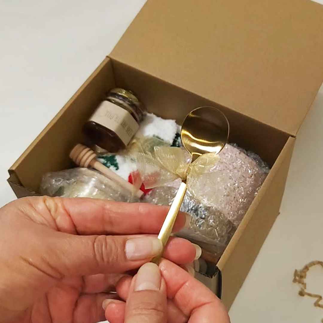 A hand holds a gold spoon with a decorative bow in front of The Cozy Sips Gift Box.