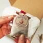 A hand holding a small clear plastic bag containing a beige fabric pouch of mulling spices, with a round paper tag reading 'Mulling Spices for cider or wine' and a cinnamon stick attached.