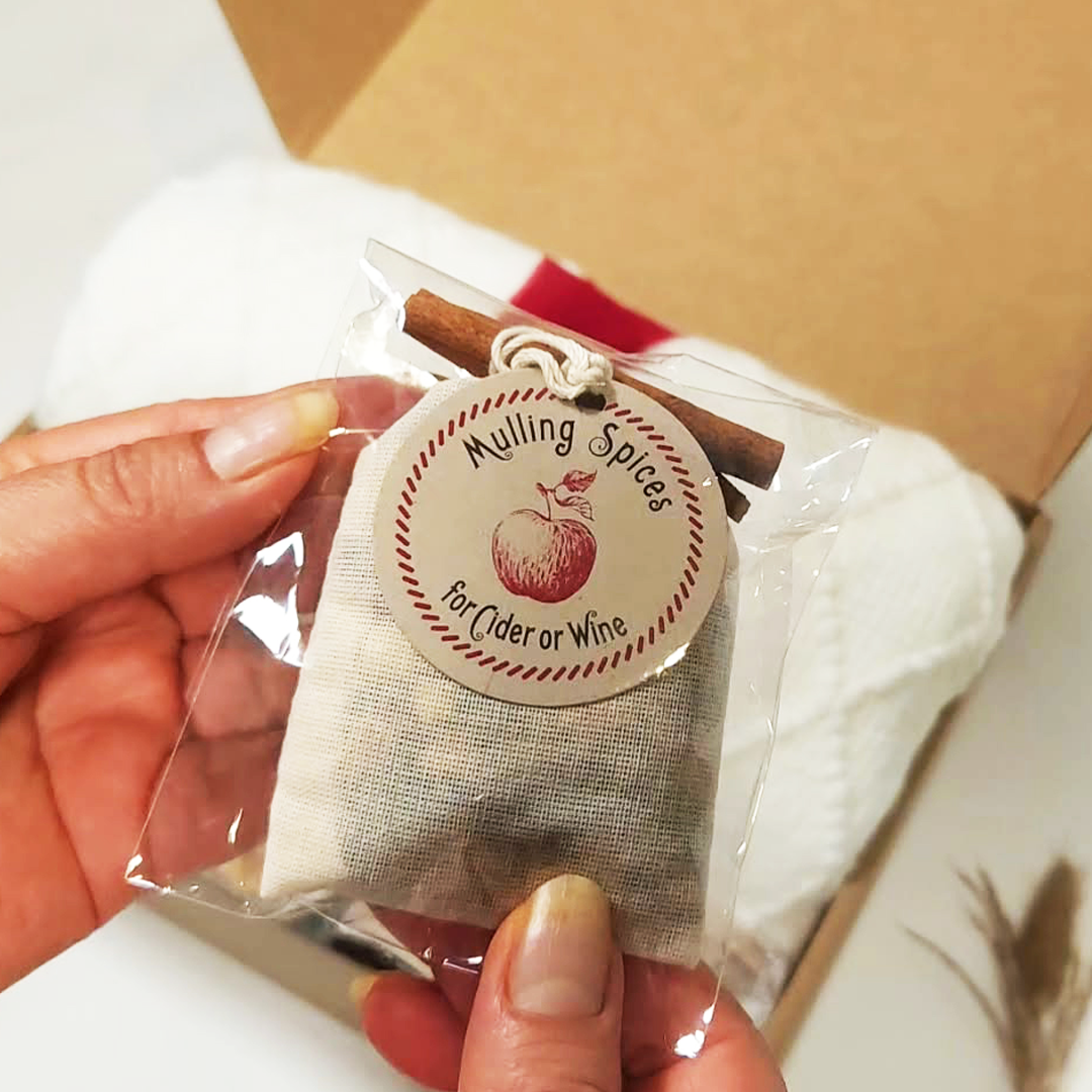 A hand holding a small clear plastic bag containing a beige fabric pouch of mulling spices, with a round paper tag reading 'Mulling Spices for cider or wine' and a cinnamon stick attached.