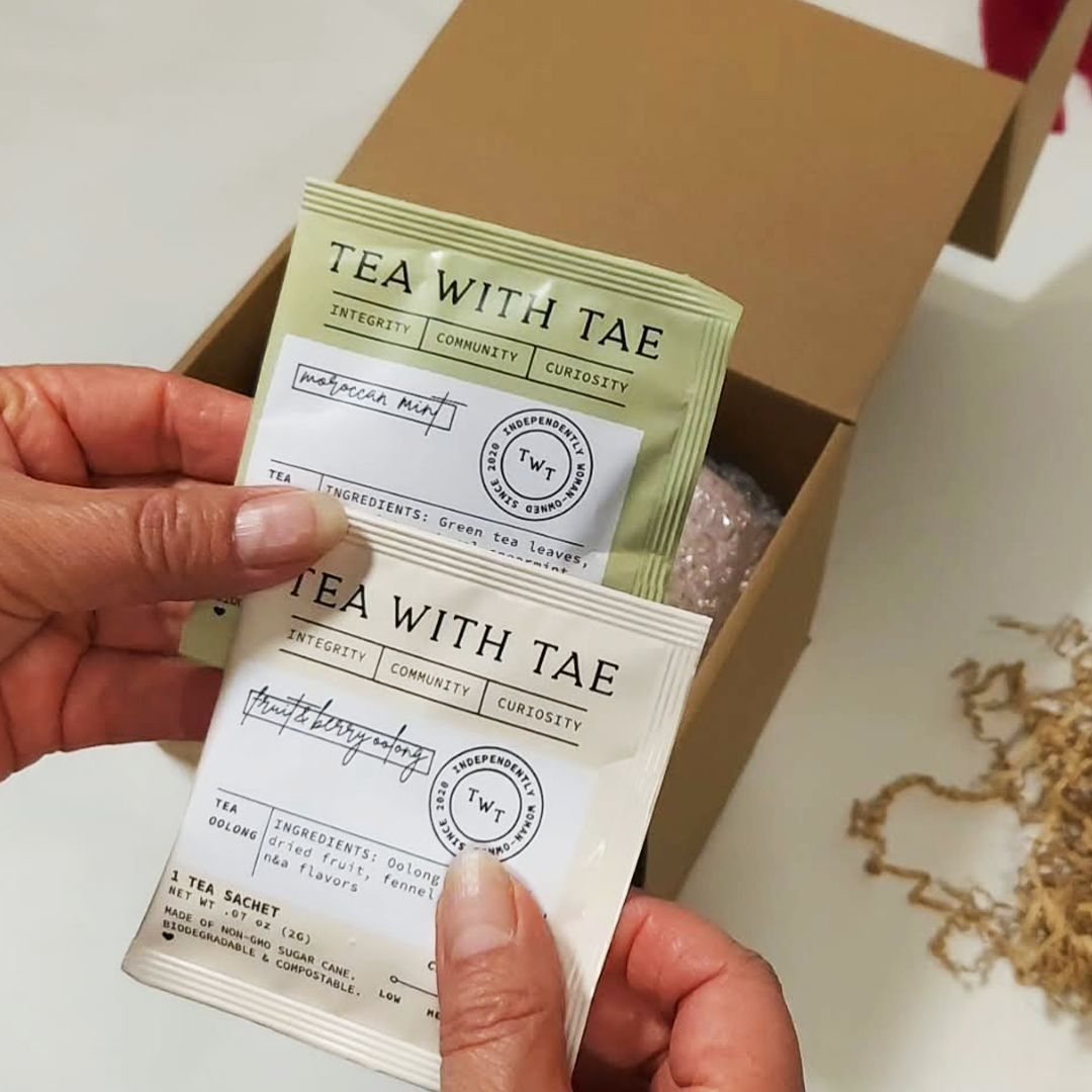 Two hands holding two 'Tea With Tae' tea sachets in front of The Cozy Sips Gift Box.