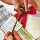 A person holds a small gold coffee or tea teaspoon over a Christmas gift box containing tea packages wrapped in clear plastic and tied with a red ribbon.
