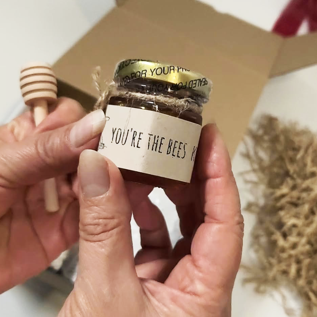A person's hands hold a small glass jar of honey with a white label that reads 'YOU'RE THE BEES. The Cozy Moments Gift Box.