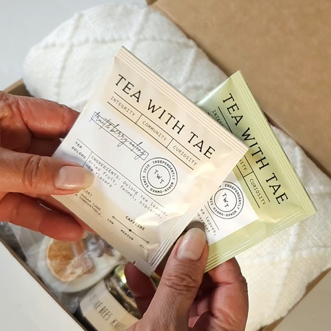 Two hands holding two individually wrapped tea bags from Tea With Tae, one in cream packaging and one in pale green packaging.