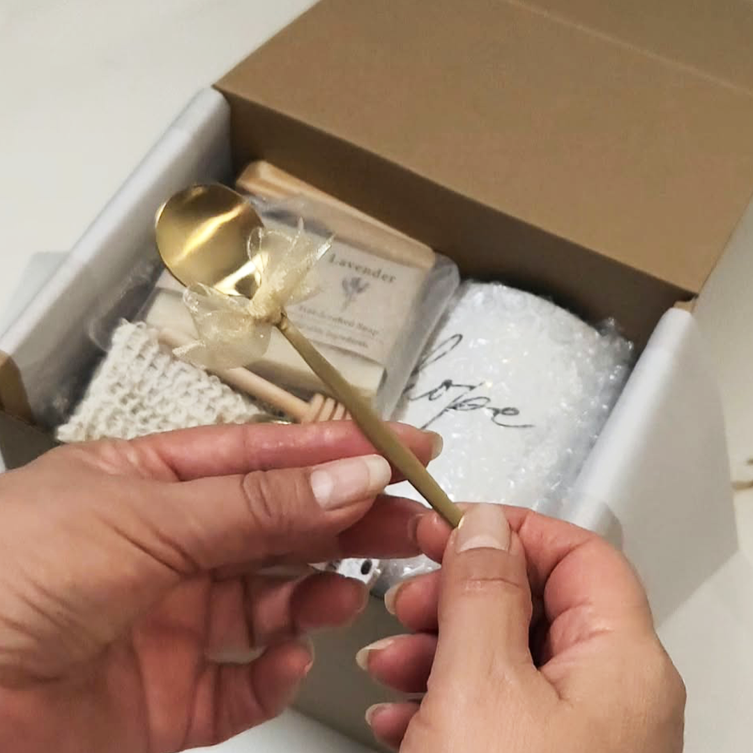A close-up of hands holding a small gold spoon with a delicate cream ribbon bow. The Cozy Moments Gift Box.