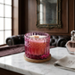 A pink gradient glass candle with a wooden wick, sitting on a cork coaster on a white marble table in a luxury room setting. Cranberry Splash-7oz posh candle vessel & lid - translucent mauve.