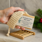 Hand holding a bar of soap with a label, placed on a wooden soap dish against a neutral background.