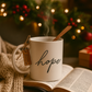 White mug with 'hope' text, steaming liquid, chocolate spoon, and gift tag on an open book with Christmas decorations in the background.