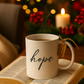 White mug with 'hope' on an open book, surrounded by festive decorations including a lit candle and blurred lights.