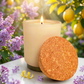 A lit cream-colored candle in a matte glass jar sits on a white marble surface, with a round cork lid leaning against the front. The scene is styled with purple flowers, a lemon peel, and a blurred background of lemons and greenery.