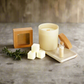 A candle set arranged on a gray stone-like surface with a wooden backdrop, including a cream-colored candle in a glass jar, three cream wax cubes, a small clear bottle with wooden wicks, a square kraft paper box, a cork stopper, and a small tray with matches. A sprig of greenery rests beside the items.