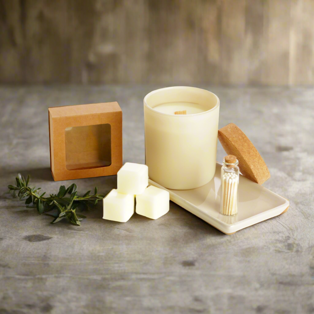 A candle set arranged on a gray stone-like surface with a wooden backdrop, including a cream-colored candle in a glass jar, three cream wax cubes, a small clear bottle with wooden wicks, a square kraft paper box, a cork stopper, and a small tray with matches. A sprig of greenery rests beside the items.