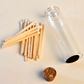 A clear glass vial with a cork stopper, shown next to a small pile of wooden matchsticks on a plain light background.
