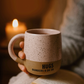 Person holding a speckled mug with 'HUGS NUMBERS 6:24-26' text in front of a candle.