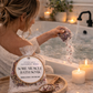 Woman in a bathtub with Epsom salt, peppermint, and rose bath soak, surrounded by candles and plants.