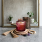 A pink glass candle jar sits on a stack of natural wood slices, surrounded by a wooden bead garland, tassel decor,  in front of a mirror. 
Velvet Rose Musk-7oz posh candle vessel & lid - translucent mauve 5