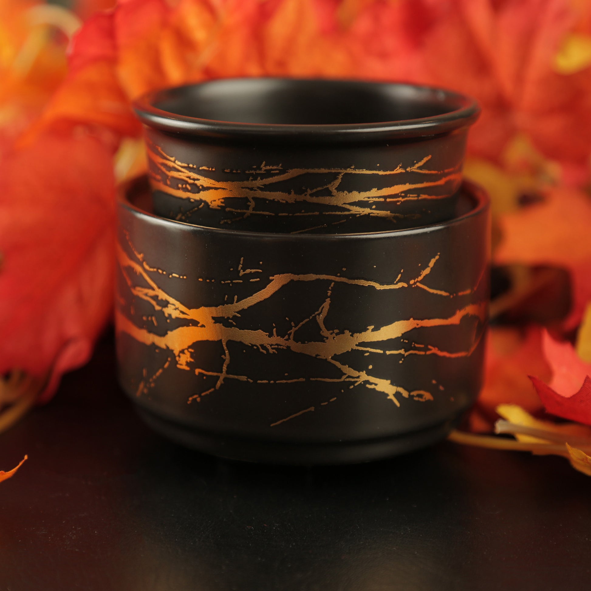 Black ceramic pot with gold branch-like patterns against a blurred autumn leaf background