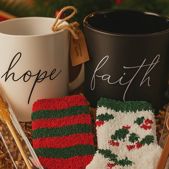 Two mugs labeled 'hope' and 'faith' with socks and chocolate and sugar stick spoons in a decorative box with a festive background.