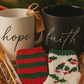 Two mugs labeled 'hope' and 'faith' with socks and chocolate and sugar stick spoons in a decorative box with a festive background.